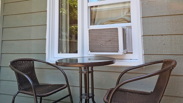 Un patio cubierto con una mesa y dos sillas de mimbre, una ventana con persianas y una pared con revestimiento horizontal.