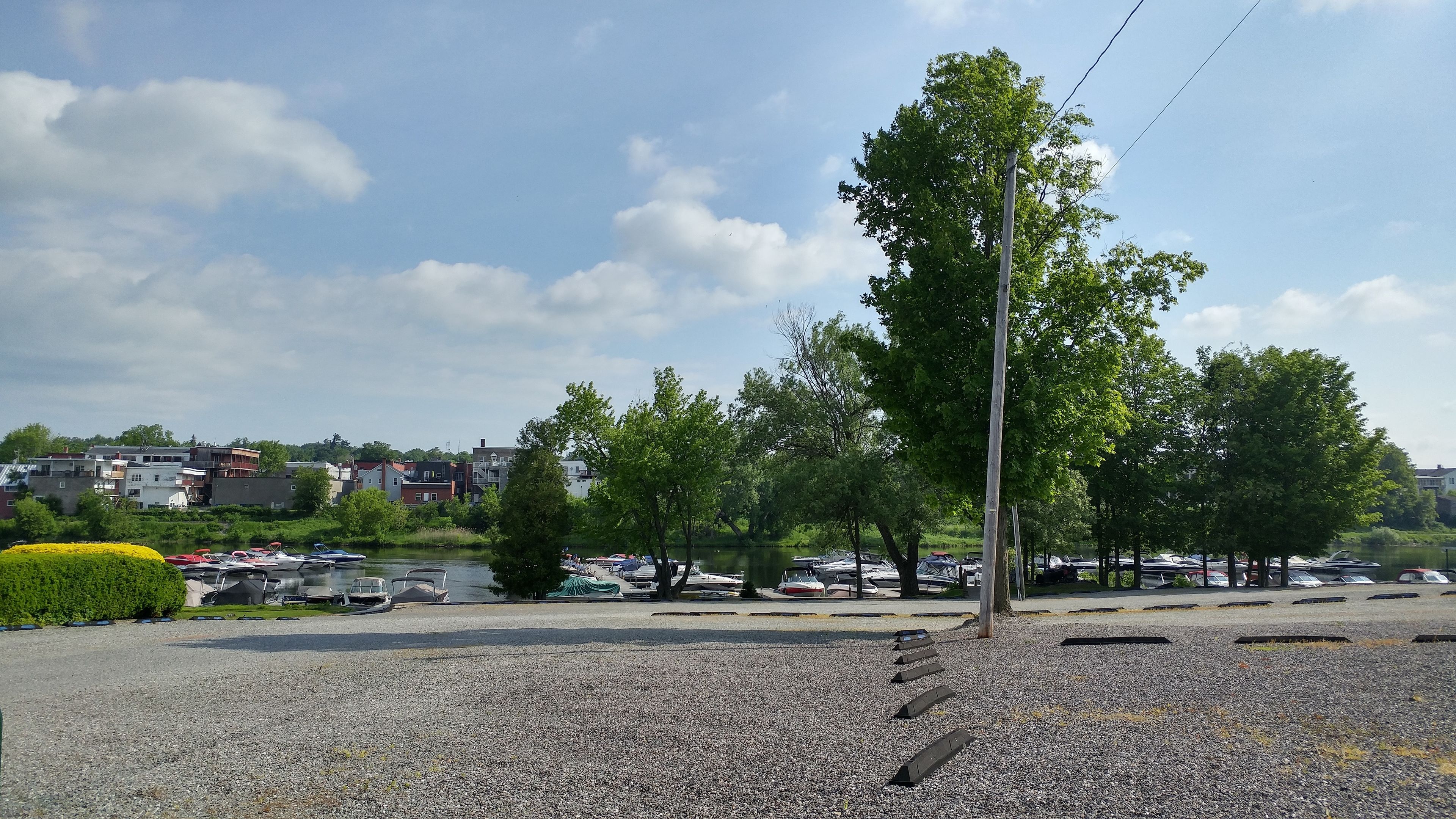 Un espace de stationnement avec un sol en gravier, des arbres et une étendue d’eau en arrière-plan.