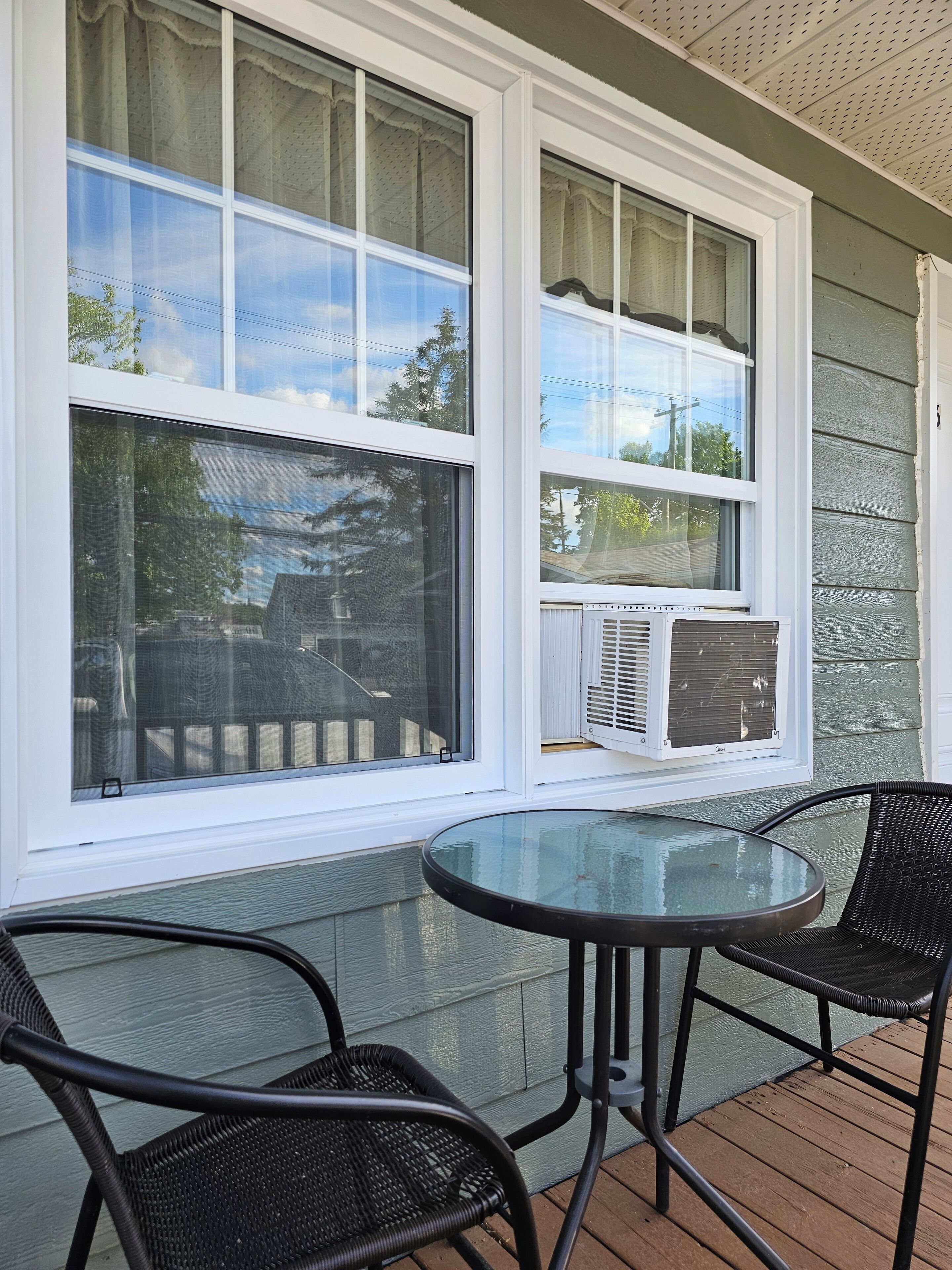 A covered outdoor seating area with a glass table and two wicker chairs, adjacent to a window with an air conditioning unit.
