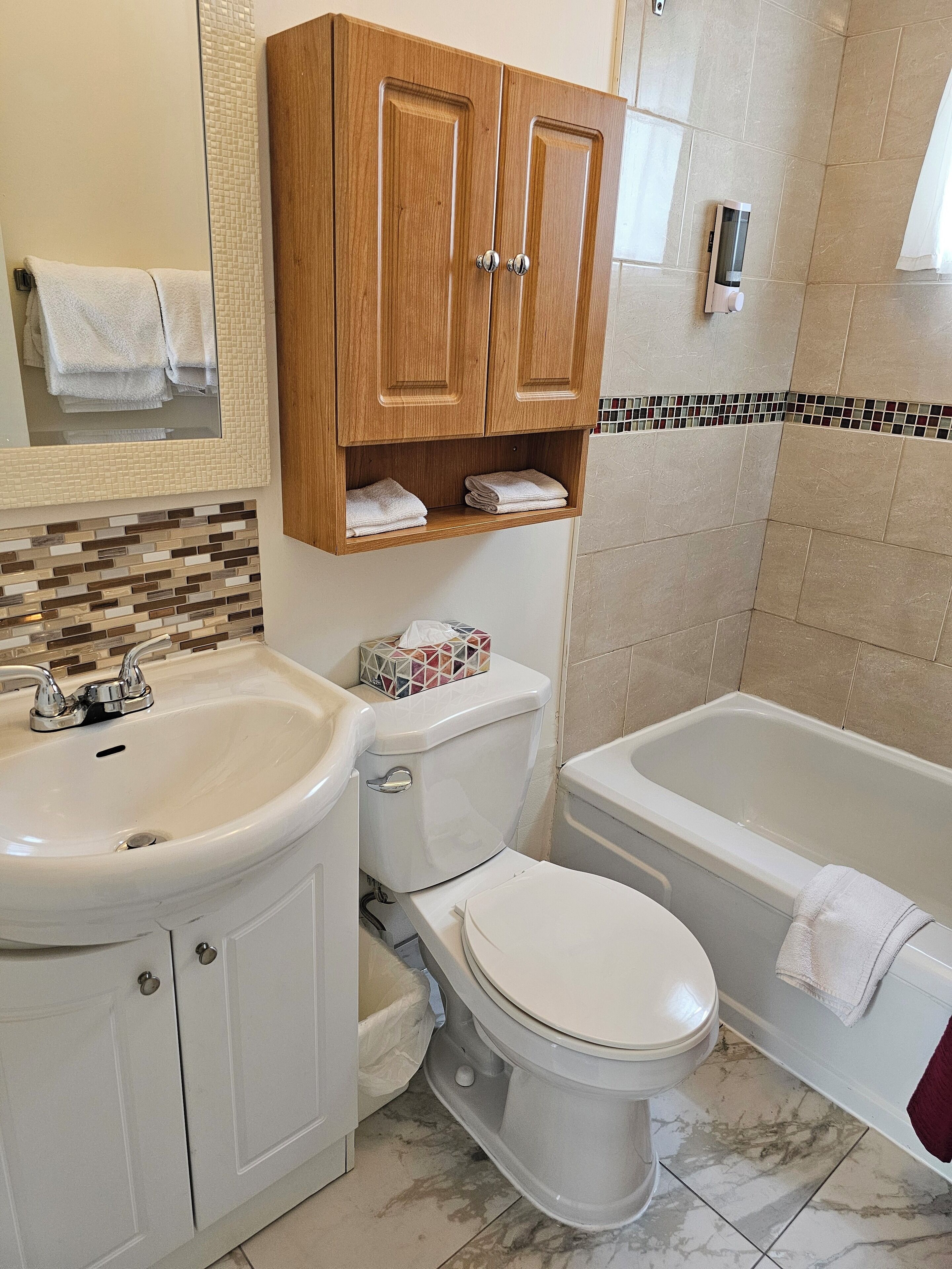 A bathroom with a sink, toilet, and bathtub. There are wooden cabinets above the toilet and a tiled backsplash above the sink.
