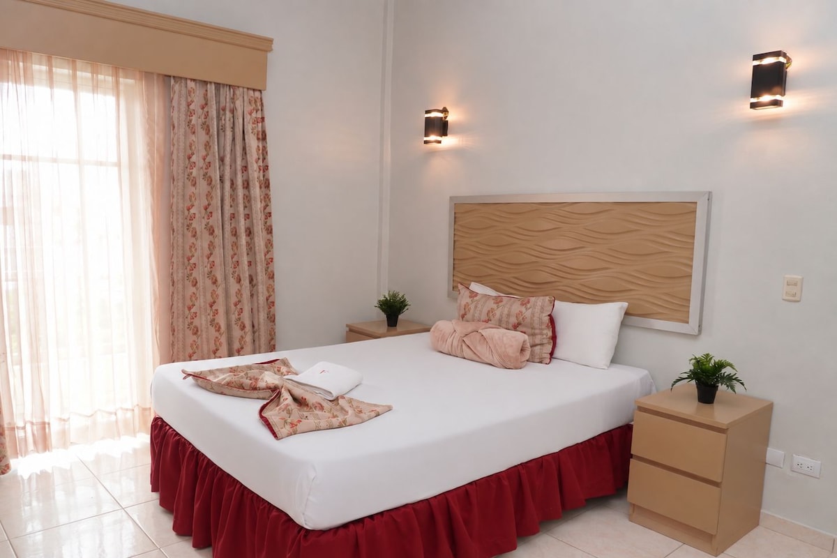 Una habitación de hotel con una cama grande, un cabecero de madera, dos mesitas de noche con lámparas, una ventana con cortinas y una planta pequeña en la mesita.
