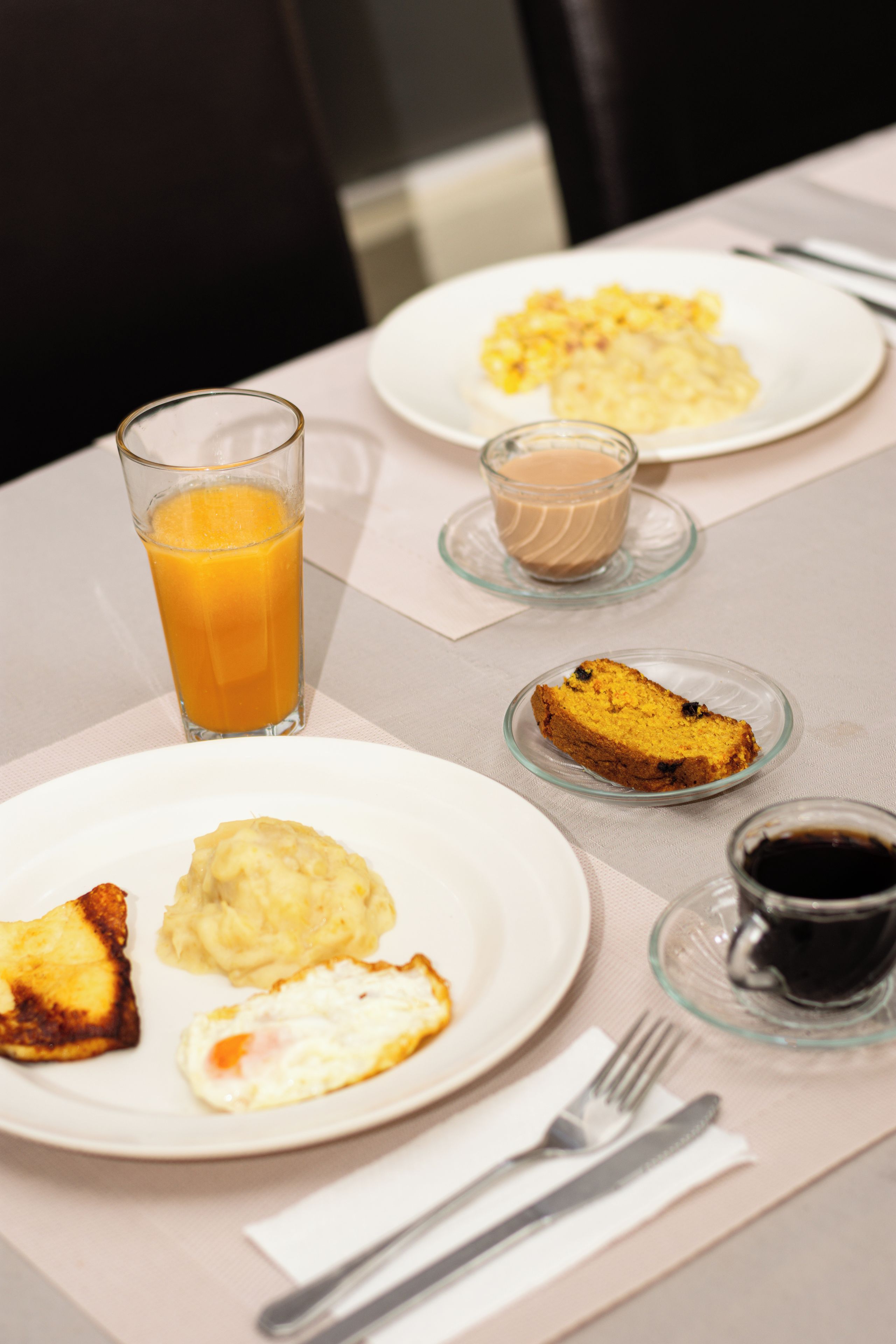 A breakfast setting with plates of food, a glass of orange juice, a cup of coffee, and a slice of cake on a table.