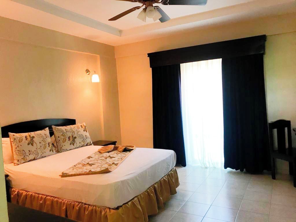 Una habitación de hotel con cama, ventilador de techo, mesita de noche y ventana con cortinas.