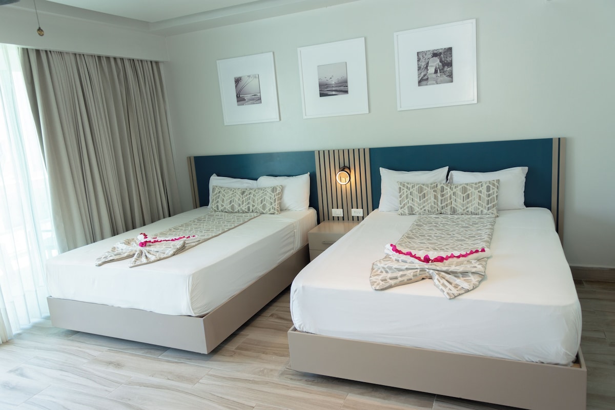 Habitación de hotel con dos camas, un ventanal con cortinas y tres cuadros en la pared.