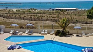 A pool area with multiple lounge chairs and umbrellas, a clear view of the beach and ocean, and a lifeguard tower in the background. - Les Rivages d'Ulysse (Le Grau-Du-Roi)