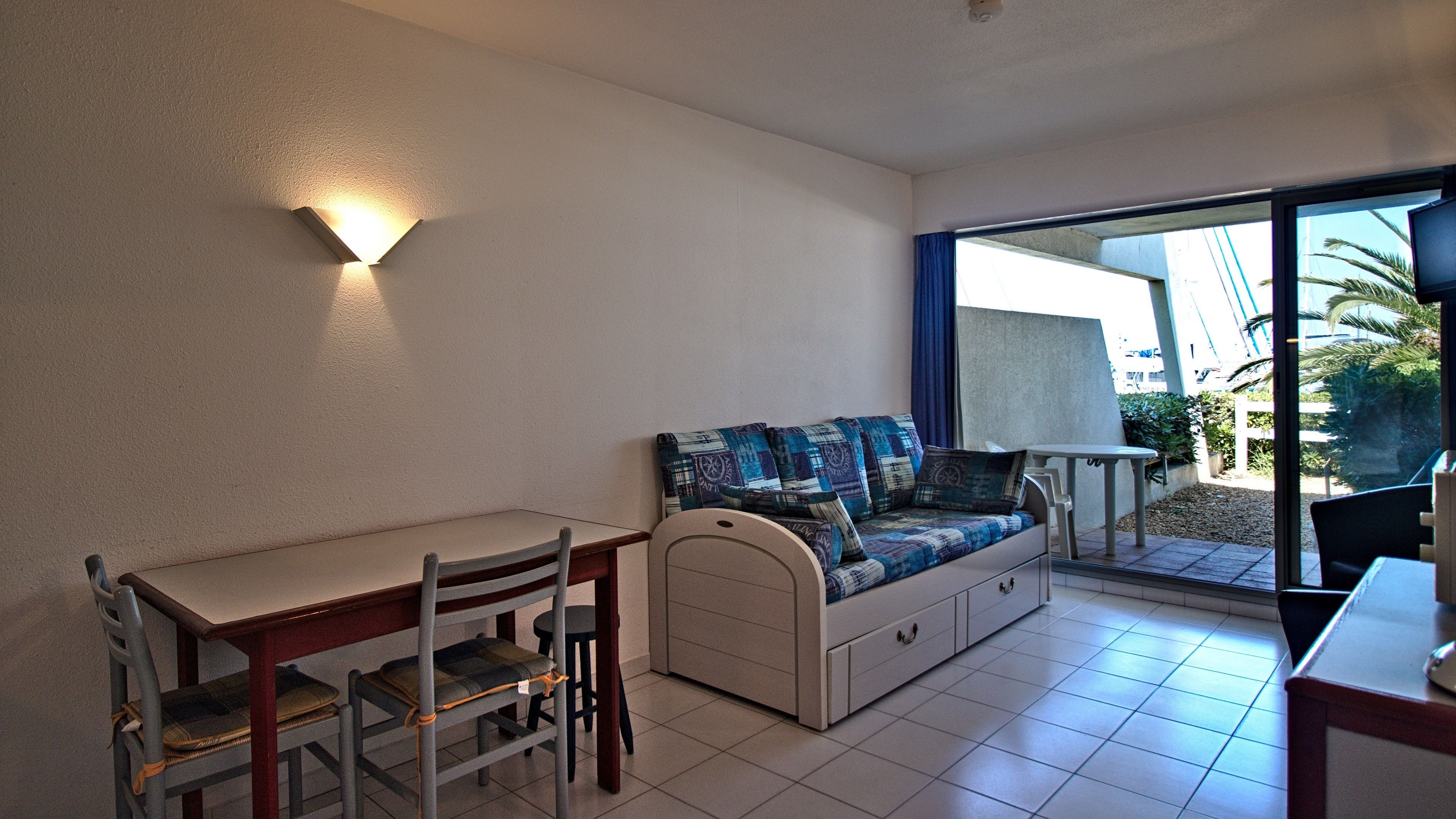 A compact living space with a dining table, chairs, a sofa with patterned cushions, a small table with a lamp, and a sliding glass door leading to an outdoor area with plants.