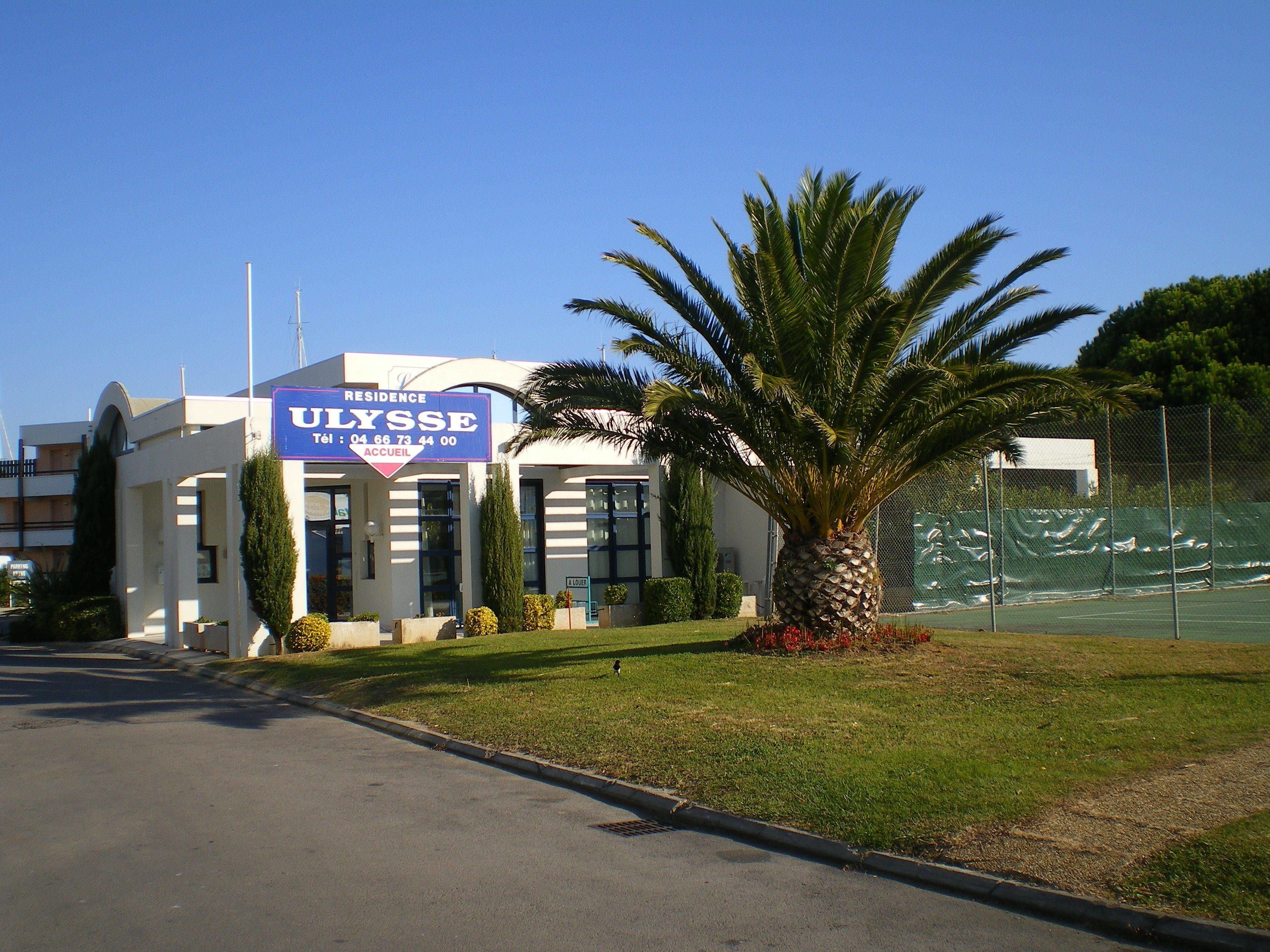 A building with a sign reading "ULYSSE RESIDENCE" and a palm tree in front.