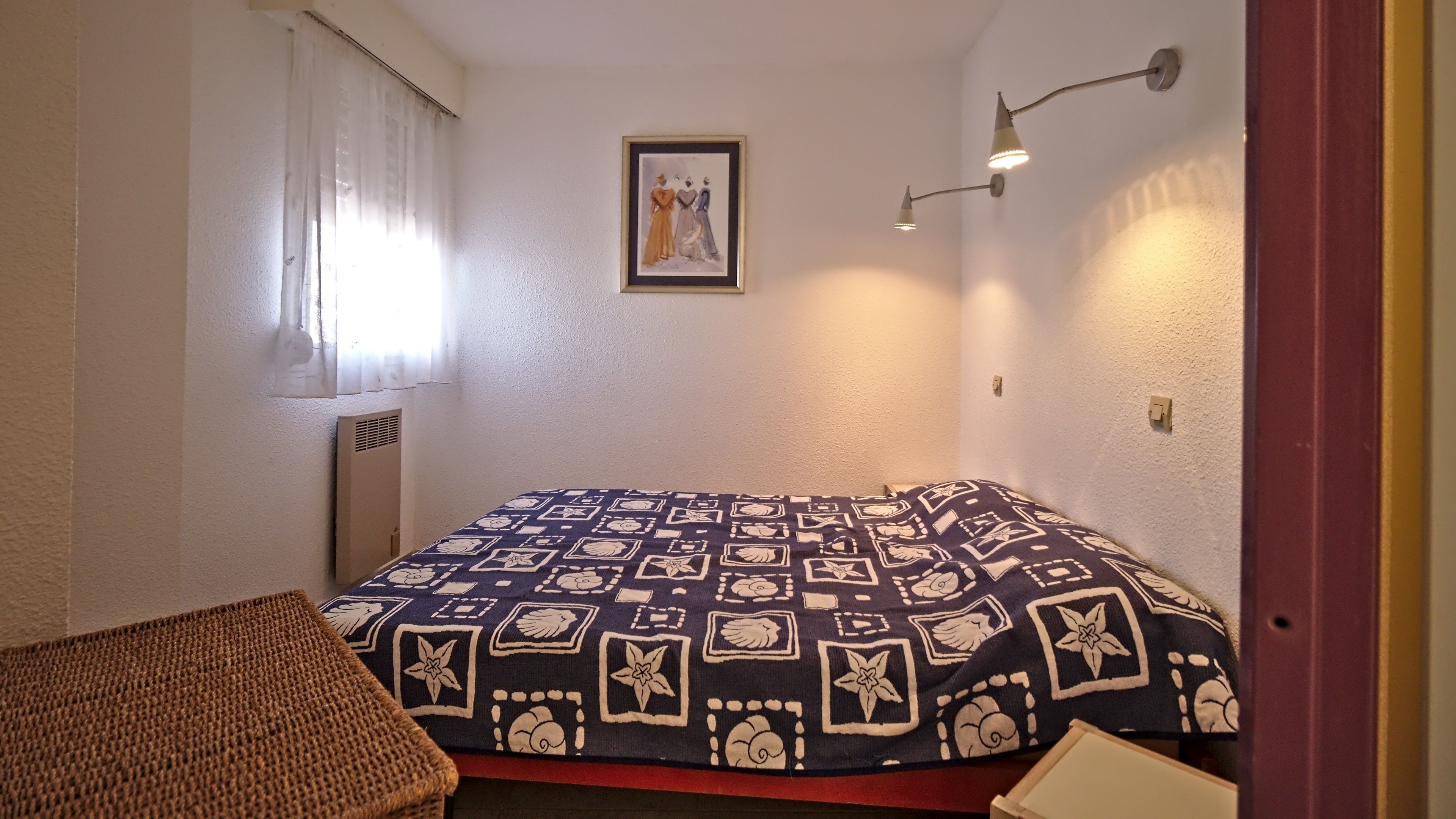 A small, neatly arranged bedroom with a single bed, a framed picture on the wall, and a wall-mounted light fixture.