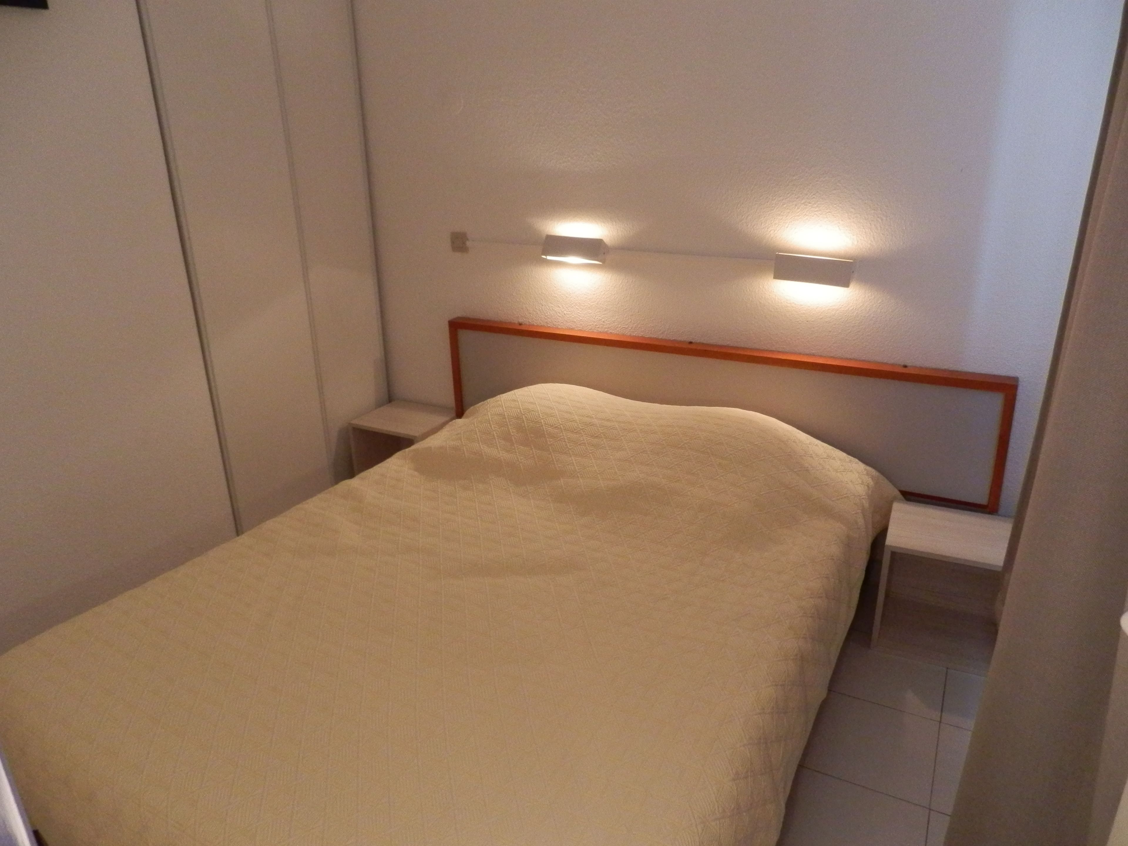 A small, neatly made bed with a wooden headboard and two wall-mounted lights.