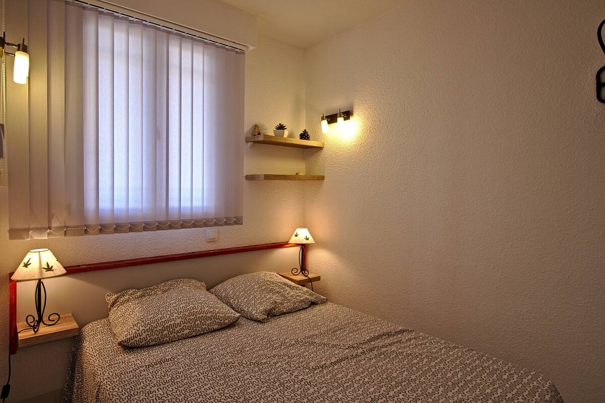 Ein Schlafzimmer mit einem Bett, zwei Nachttischlampen, einem Fenster mit Jalousien und zwei Wandregalen mit Dekoration.