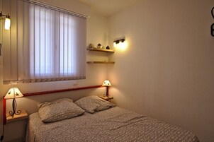 A bedroom with a bed, two bedside lamps, a window with blinds, and two wall-mounted shelves with decorative items.
