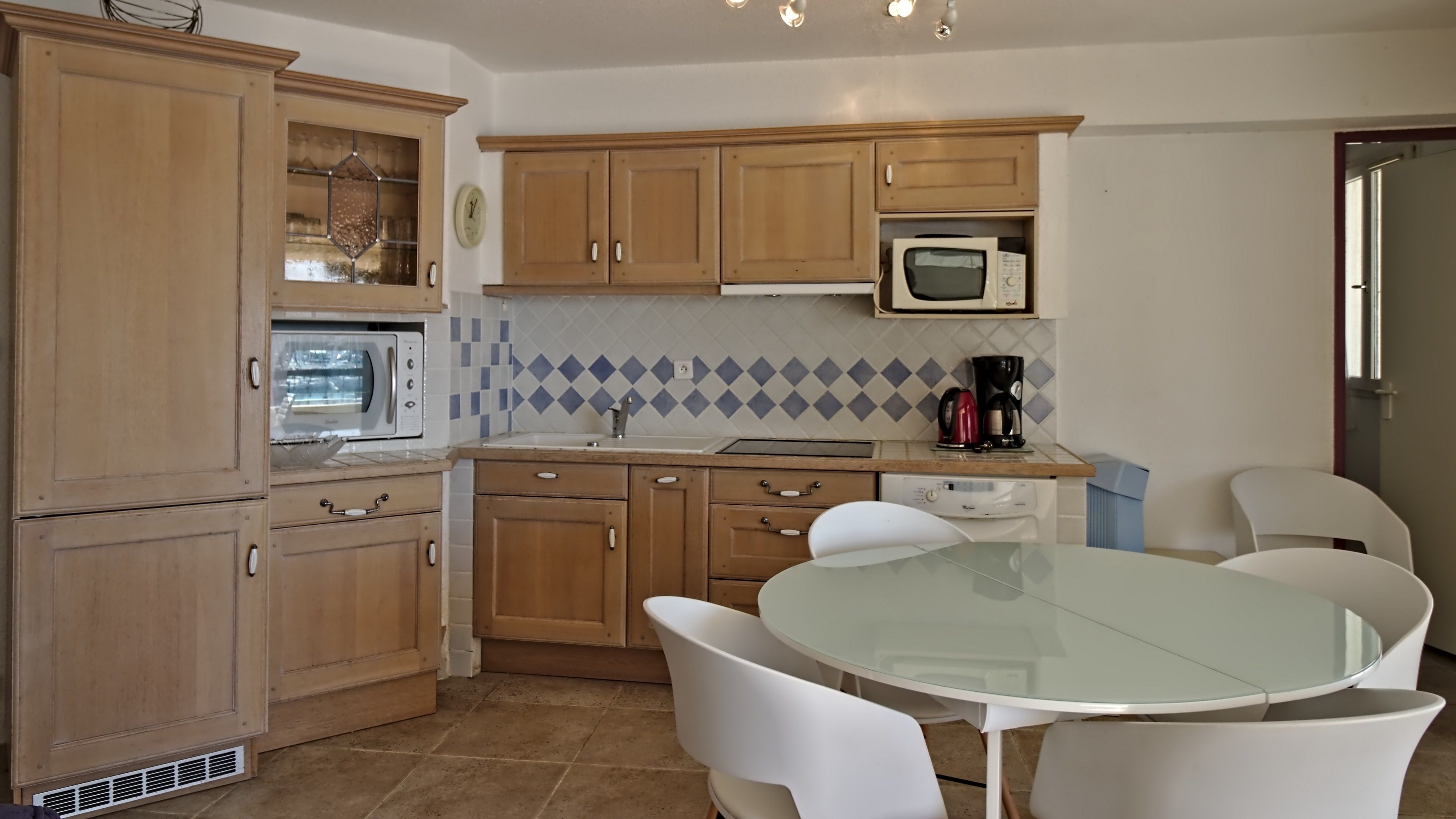 A kitchen with wooden cabinets, a microwave, a coffee maker, and a round glass table with white chairs.
