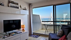 A modern living room with a TV, books, and a lamp. A balcony with a view of the marina is visible. - Les Rivages d'Ulysse (Le Grau-Du-Roi)