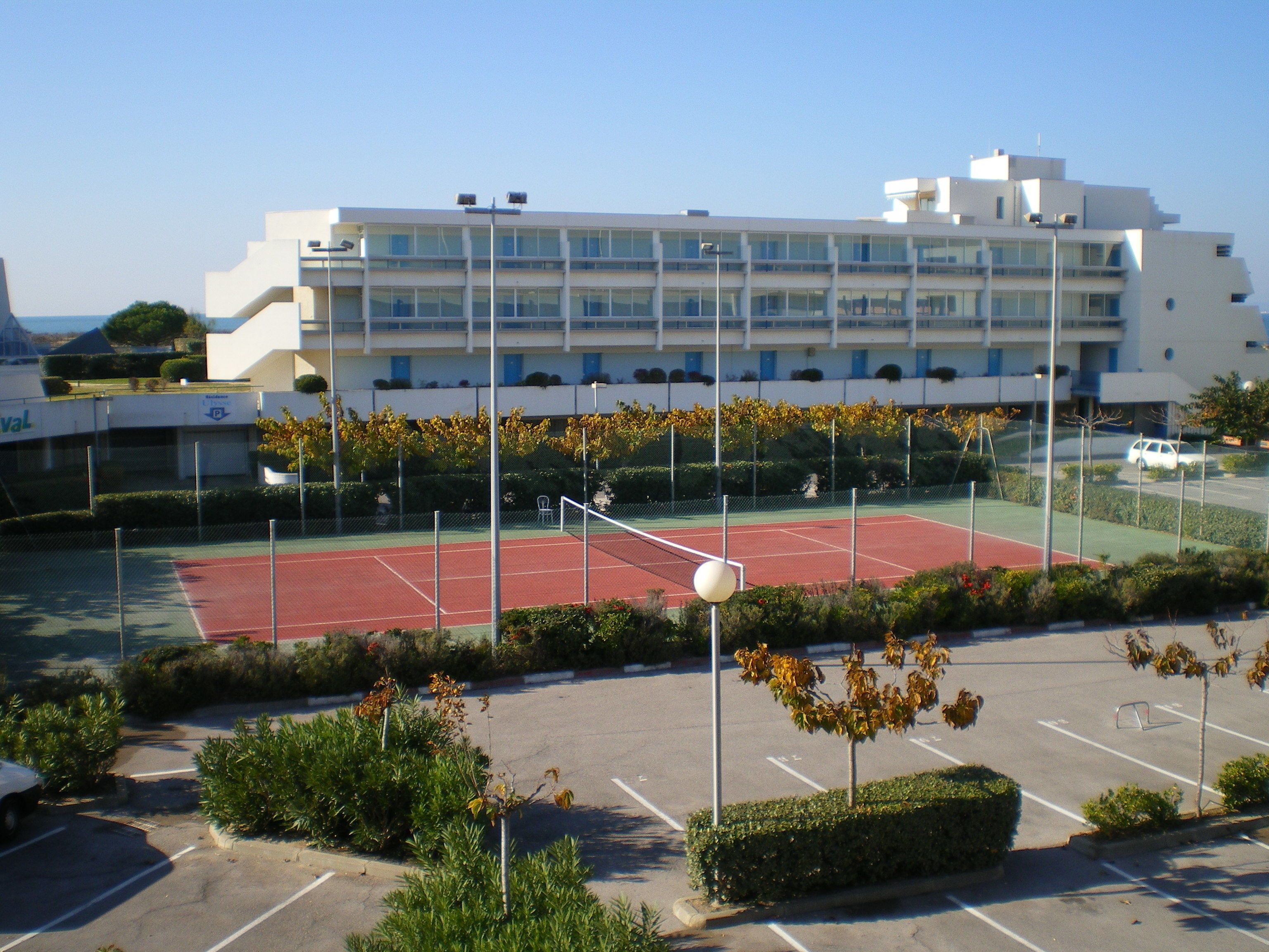 A tennis court with a net, surrounded by a parking lot and a building with multiple floors and balconies.