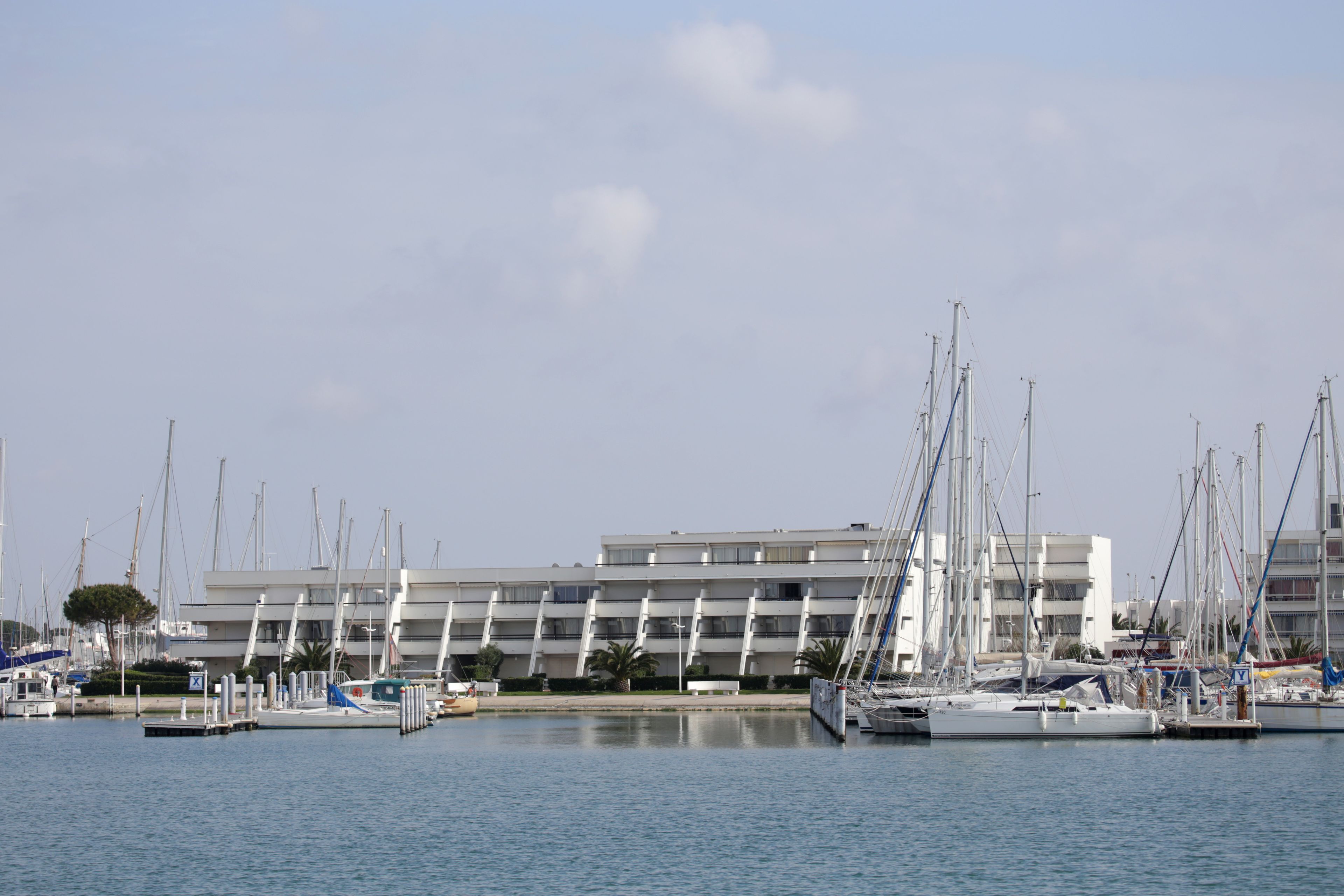 Una marina con diverse barche a vela ormeggiate, un edificio moderno con balconi e acque calme.