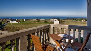 A balcony with a table and two chairs, overlooking a coastal landscape with houses and fields.