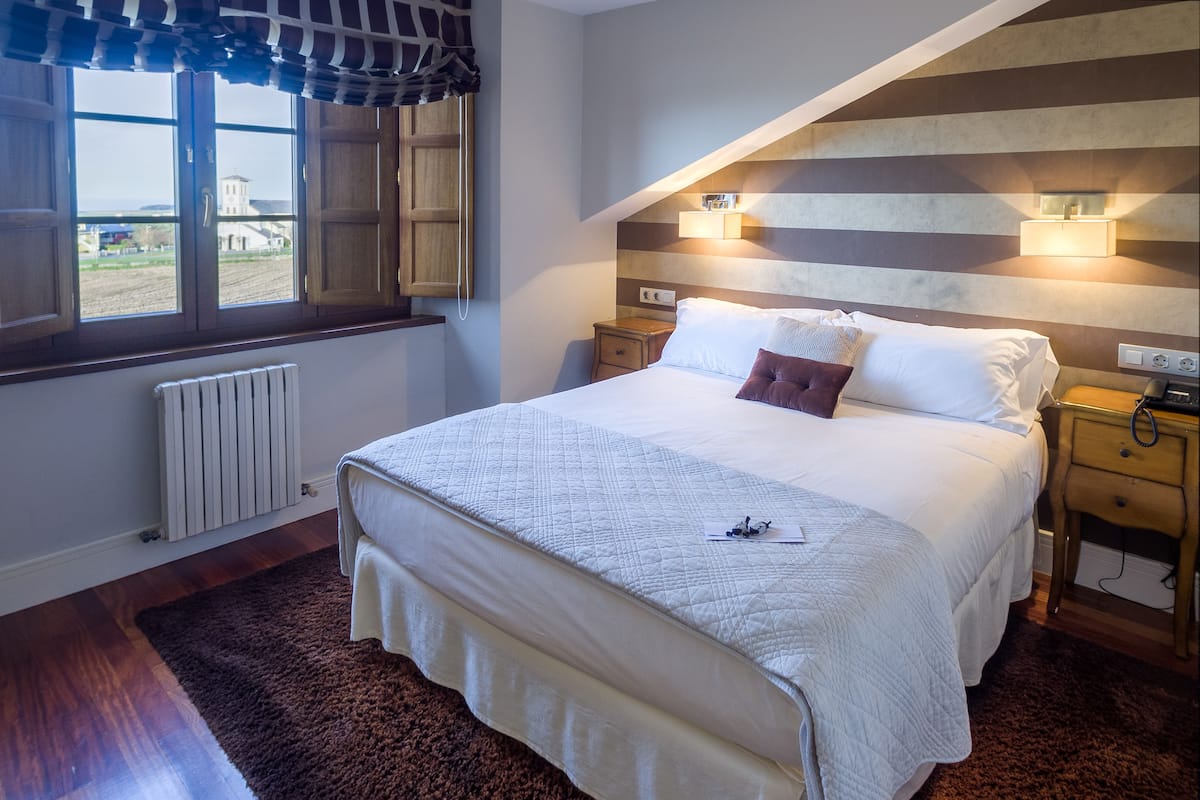 Ein ordentlich bezogenes Bett mit einer Steppdecke, ein Fenster mit Vorhängen, ein Nachttisch mit Lampe und eine Heizung.