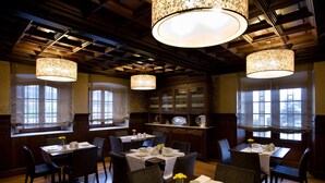 A dining area with wooden tables and chairs, chandeliers, and a view of the outdoors through large windows.