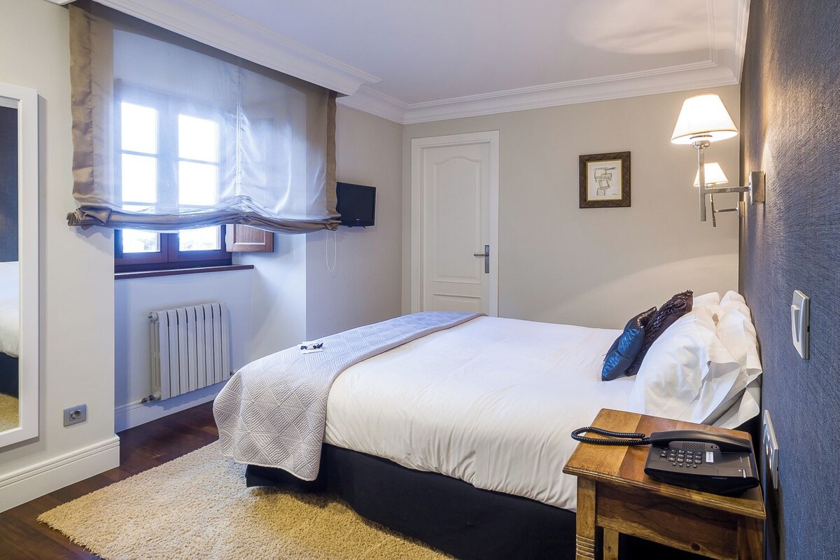A bedroom with a bed, a bedside table with a phone, a radiator, a window with curtains, and a wall-mounted TV.