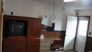 Ein Zimmer mit einem Holzkasten, einem Fernseher, einem Schreibtisch, einem Stuhl und einer Tür.