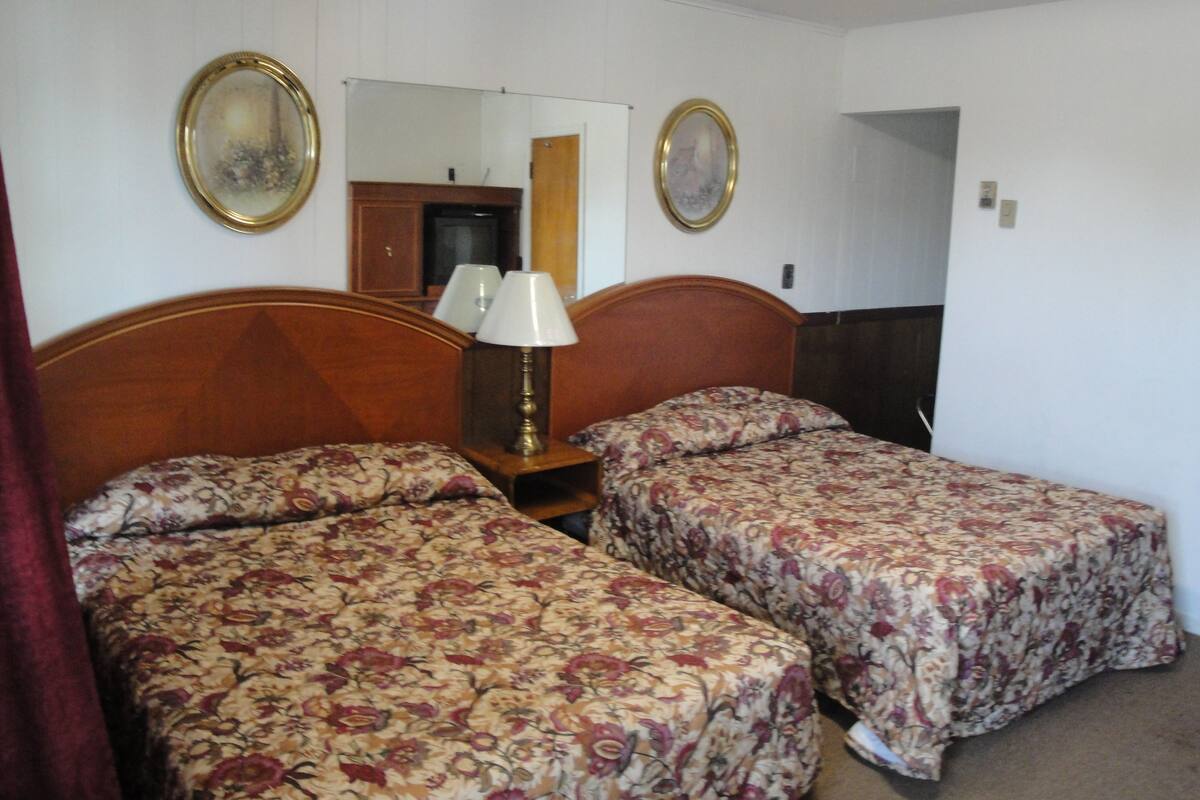 A hotel room with two single beds, a wooden headboard, a nightstand with a lamp, and two framed pictures on the wall.