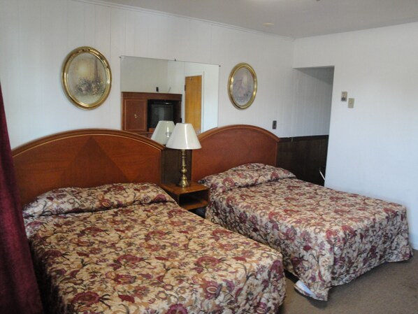 A hotel room with two single beds, a wooden headboard, a nightstand with a lamp, and two framed pictures on the wall.