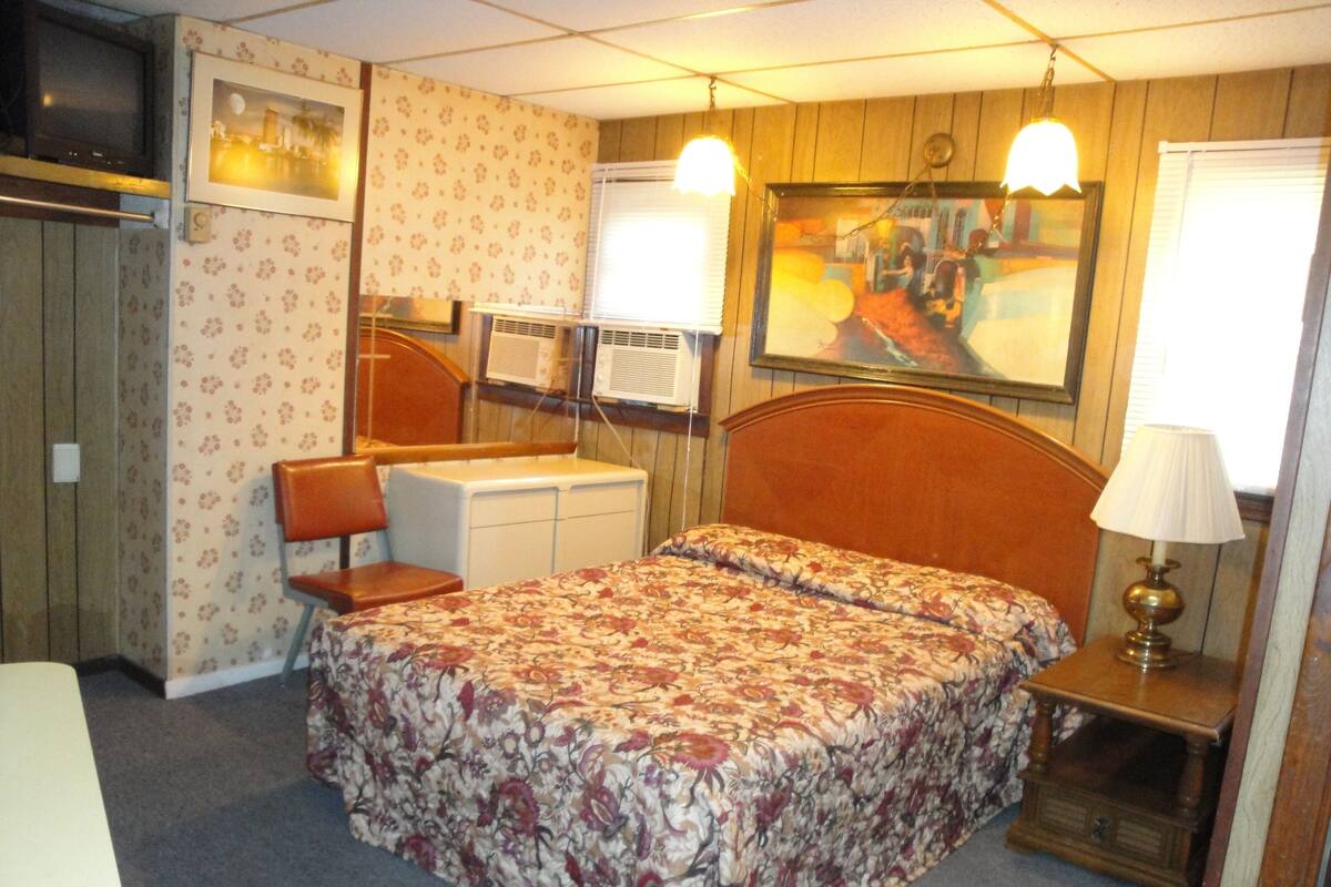 A bedroom with a bed, a desk, a chair, a lamp, and a painting on the wall.
