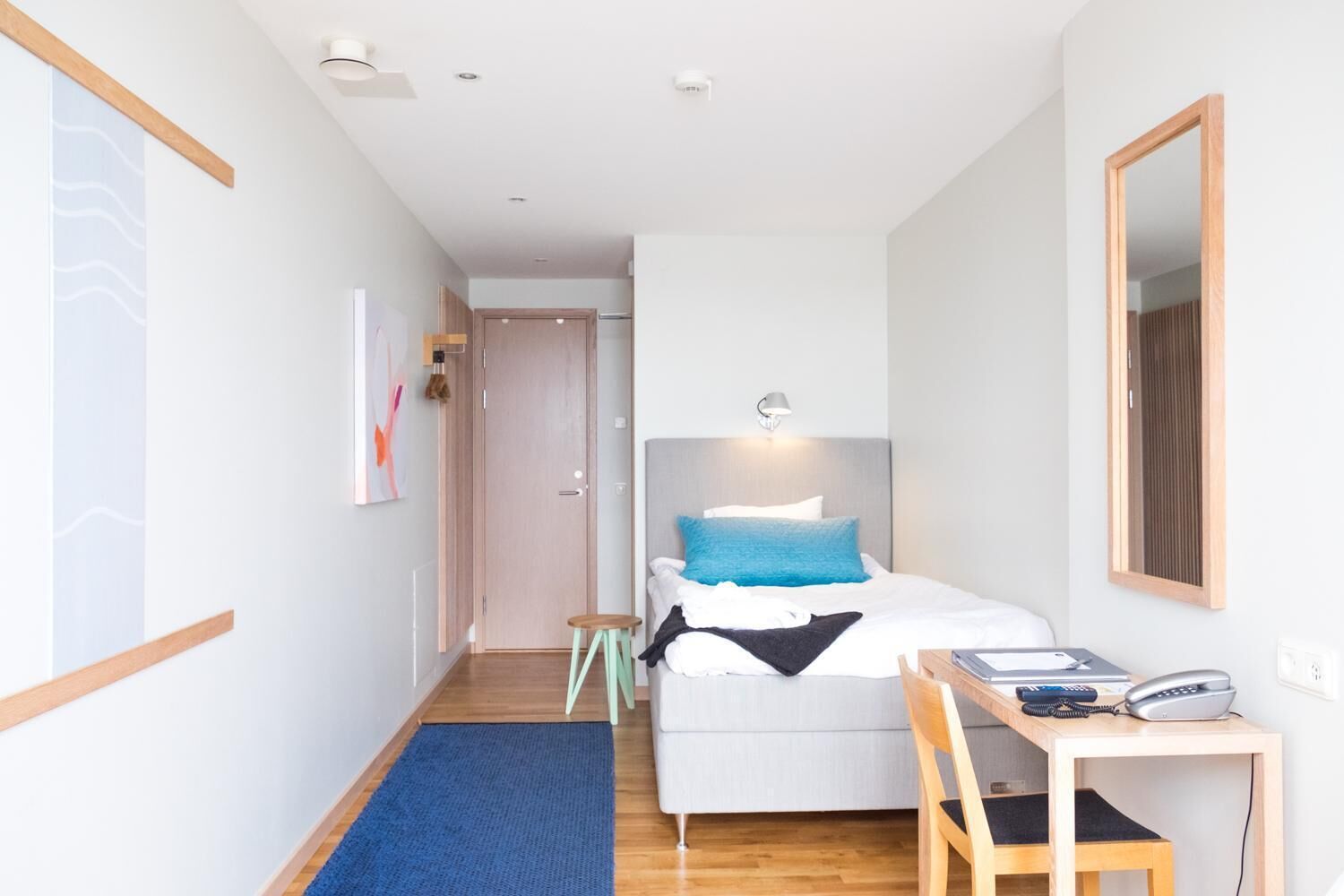 A modern hotel room with a bed, bedside table, chair, and a desk with a telephone.