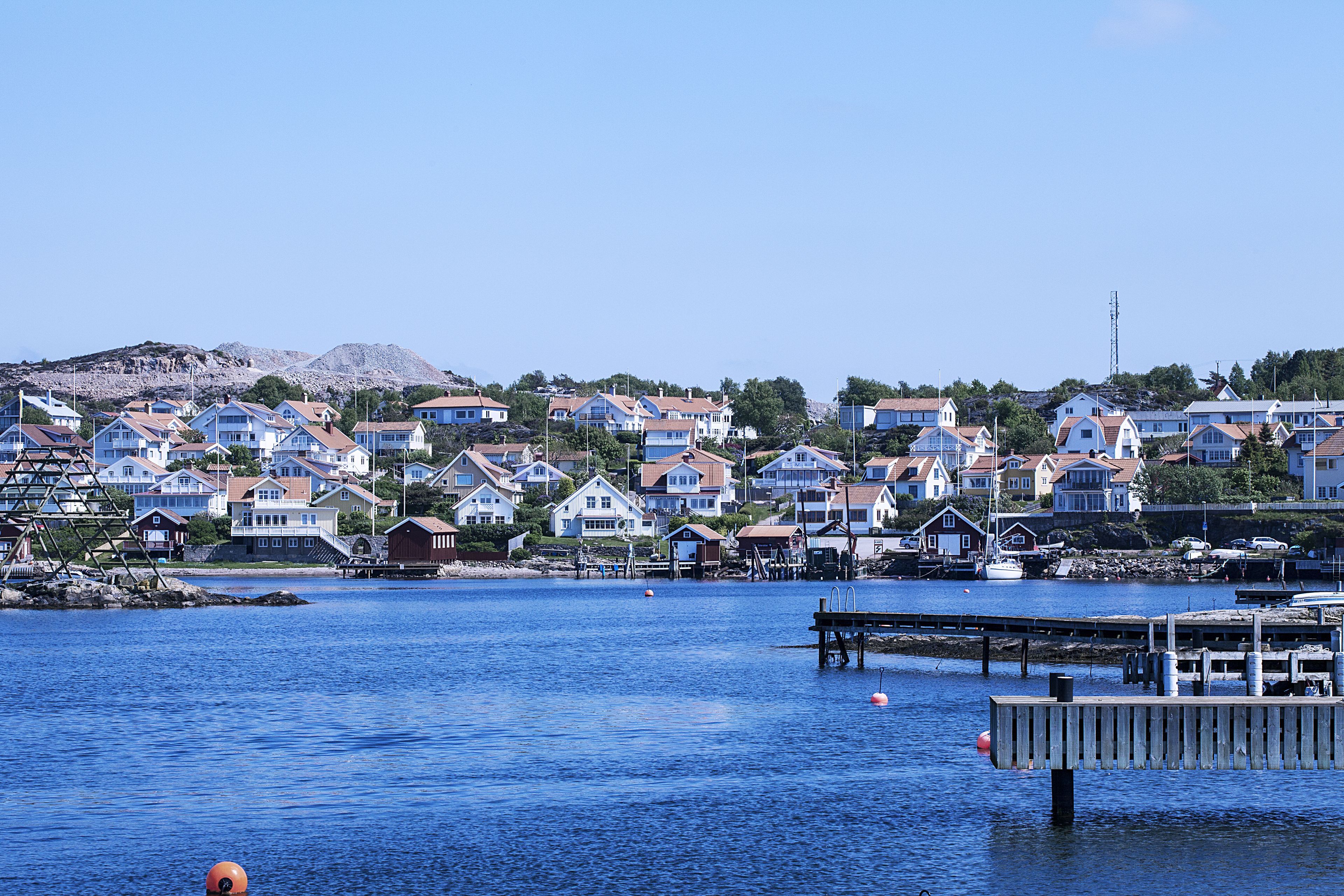 En kuststad med hus längs vattnet, en brygga och en fyr i bakgrunden.