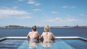 Twee personen zitten aan de rand van een overloopzwembad met uitzicht op zee en een verre skyline.