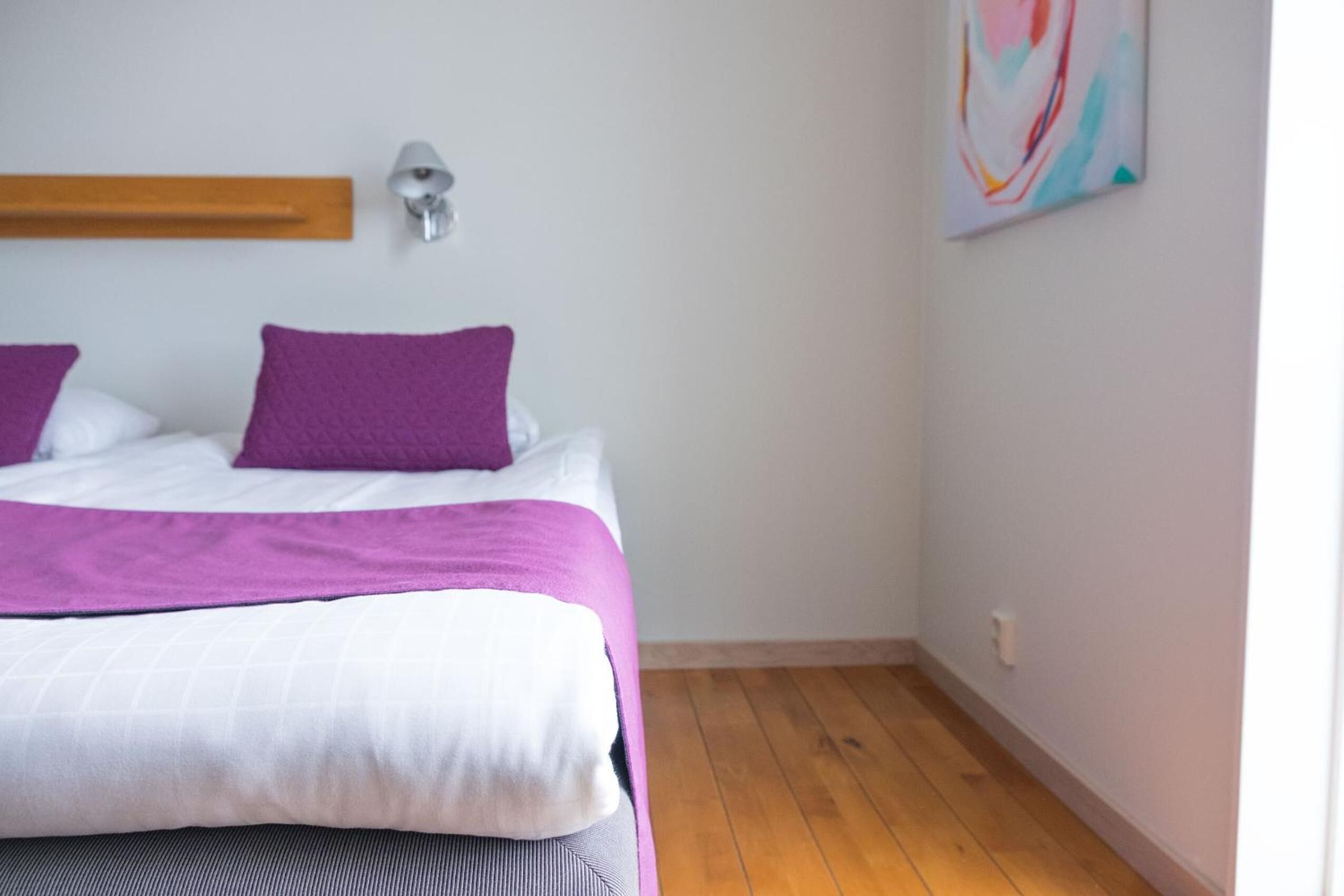 Un dormitorio con cama, almohadas moradas, colcha blanca y morada, cabecera de madera, lámpara de pared y un cuadro abstracto colorido.