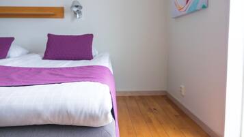 Un dormitorio con cama, almohadas moradas, colcha blanca y morada, cabecera de madera, lámpara de pared y un cuadro abstracto colorido.