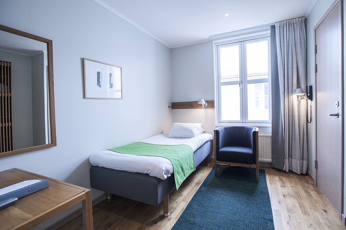 Ein Hotelzimmer mit Bett, Schreibtisch, Stuhl, einem Fenster mit Vorhängen und einem an der Wand befestigten Telefon.