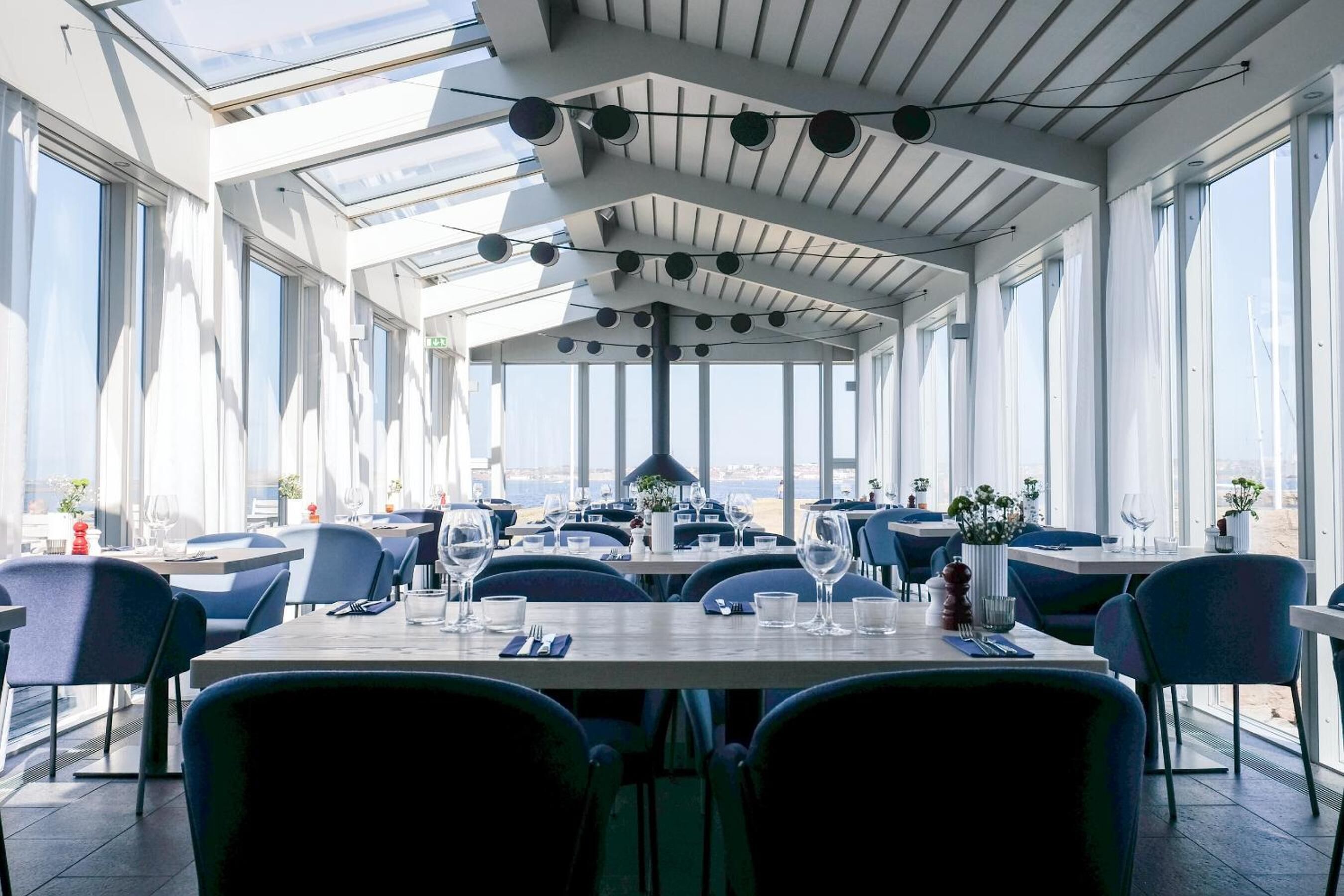 Un restaurante con techos altos, amplios ventanales y vista al mar. Las mesas están dispuestas con copas de vino y cubiertos.