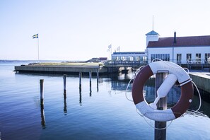 Une bouée de sauvetage est attachée à un poteau en bois, le tout situé au bord de l’eau, dans un endroit calme, avec un bâtiment visible au loin.