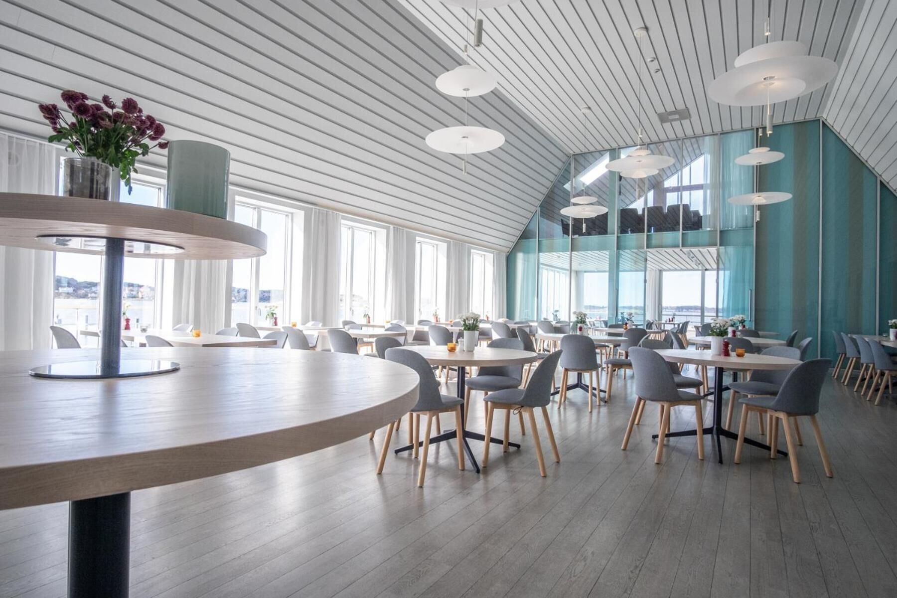 Un'ampia sala da pranzo con tavoli e sedie rotonde, grandi finestre e un soffitto alto con travi bianche.