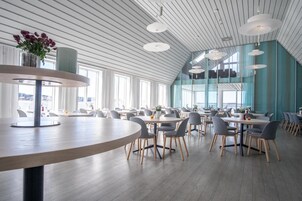 A spacious dining area with round tables and chairs, large windows, and a high ceiling with white beams.