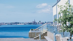 Uitzicht op het water met een houten terras, een gebouw met een blauwe balustrade en een verre skyline, inclusief een kerktoren.