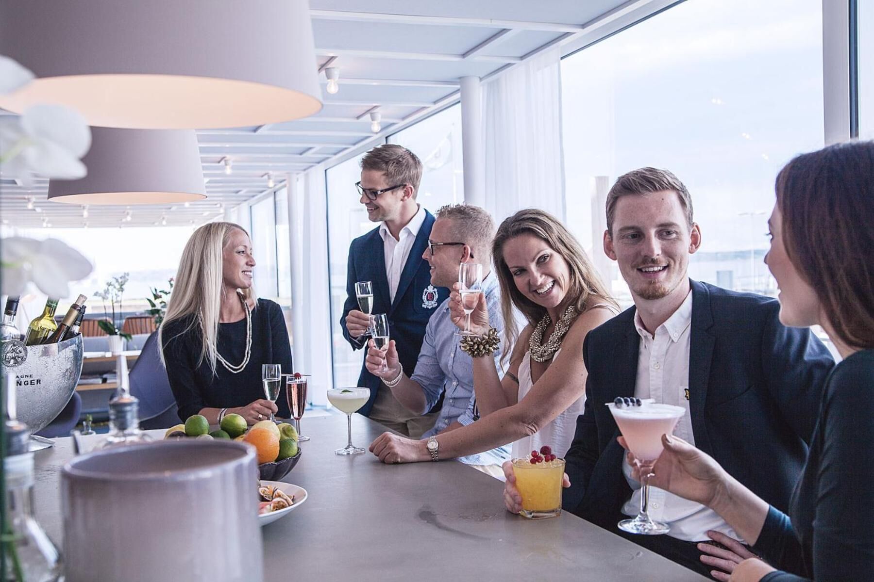 A group of people at a bar with a view of the city, bottles of alcohol, and a fruit bowl on the table.