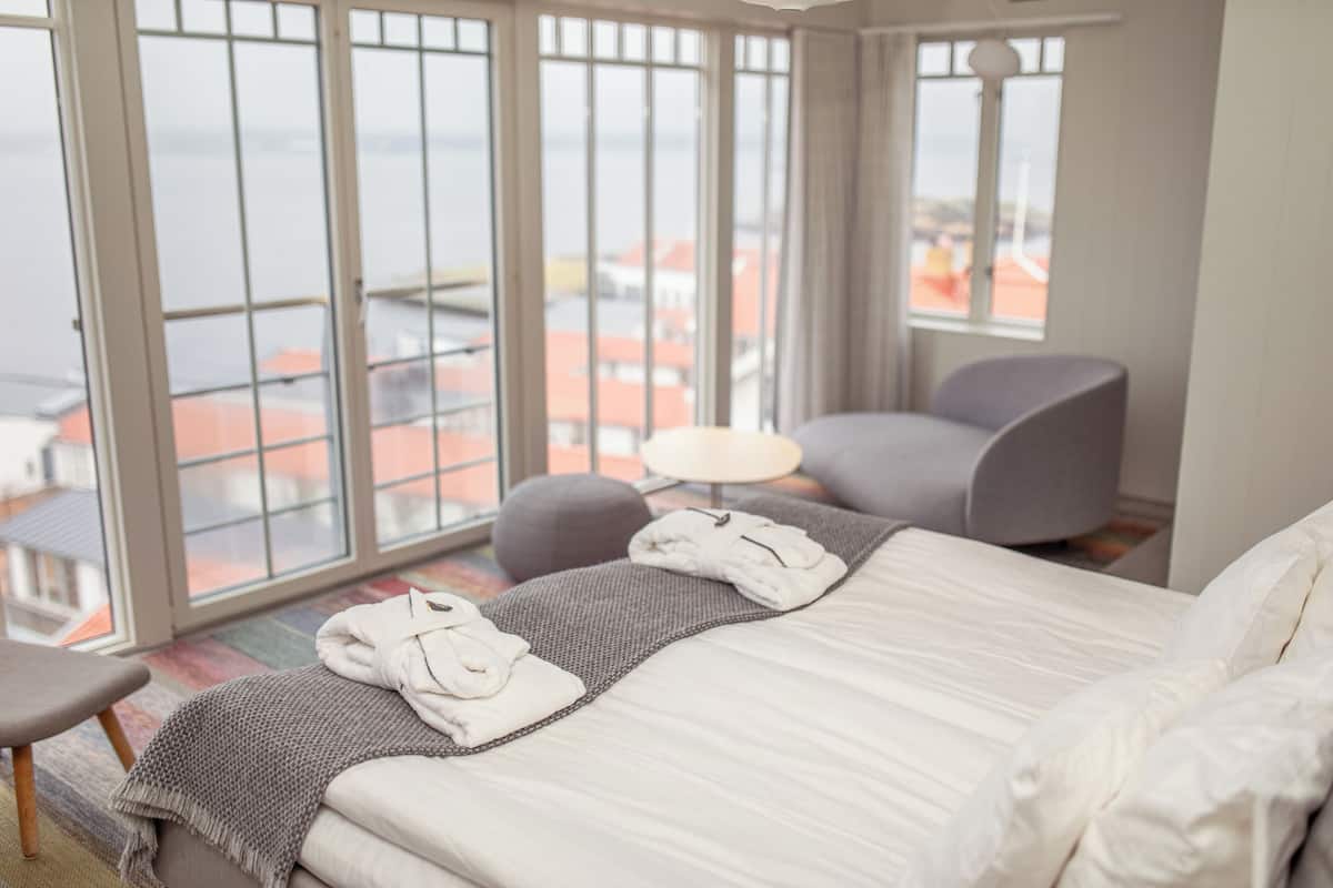 Ein Hotelzimmer mit einem großen Bett, einem Sessel, einem kleinen Tisch und Blick auf die Stadt durch große Fenster.