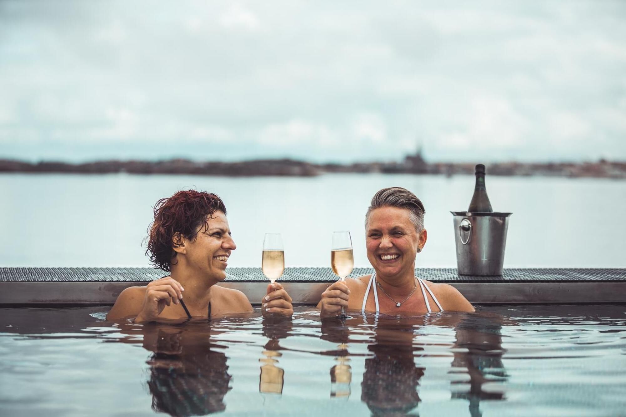 Dos personas en un jacuzzi con vista a un cuerpo de agua y una costa lejana.