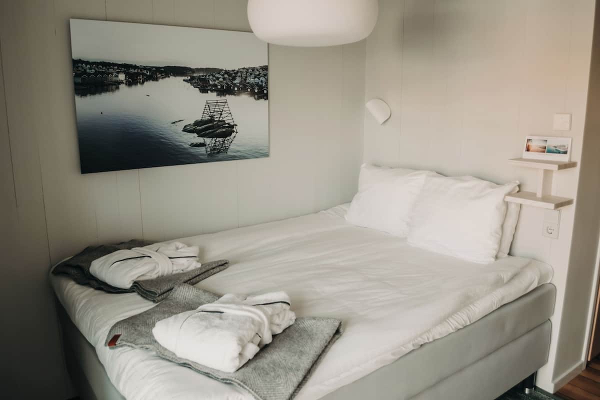 Ein ordentlich bezogenes Bett mit weißen Leinen, einer grauen Tagesdecke und einem gefalteten Handtuch. Darüber hängt ein gerahmtes Bild mit einer Küstenlandschaft.