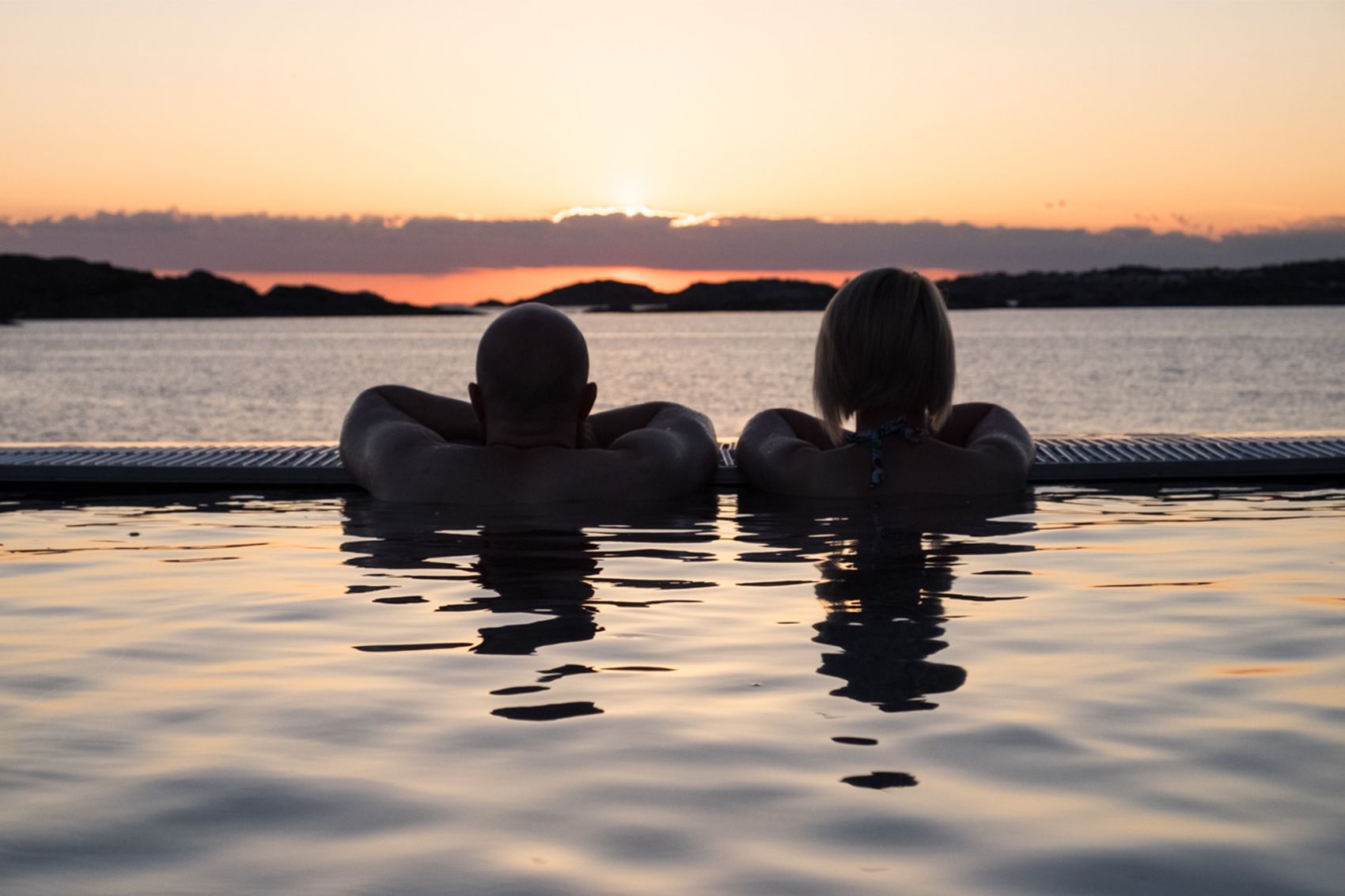 Deux personnes assises dos à dos dans une piscine, avec une vue sur le coucher de soleil.