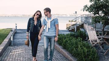 A couple walking on a paved waterfront promenade with a view of the sea and a bridge in the background.