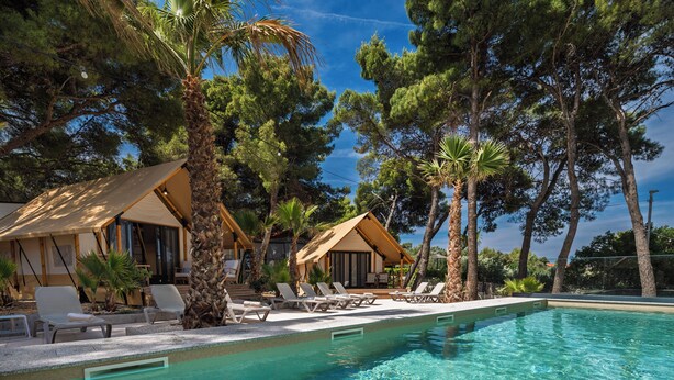 Zona de piscina con sillones de descanso y dos edificios pequeños, rodeada de árboles.