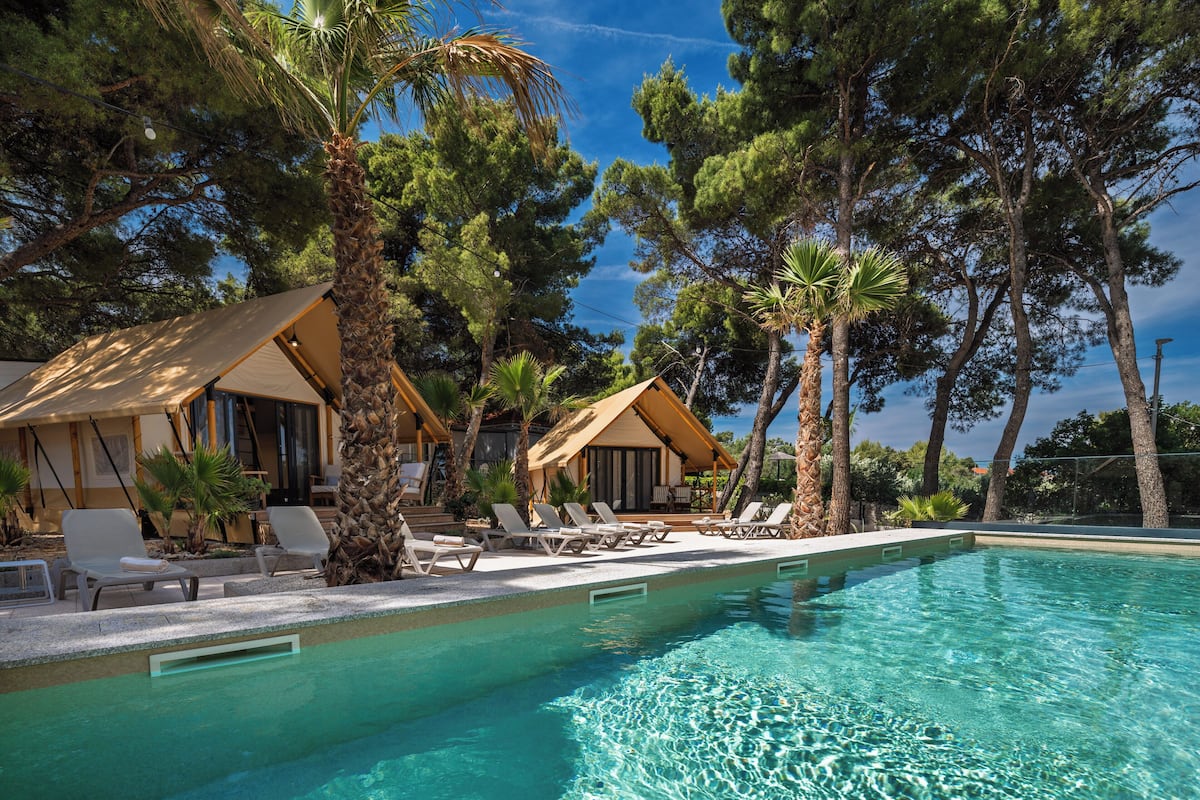 A pool area with lounge chairs and two small buildings, surrounded by trees.