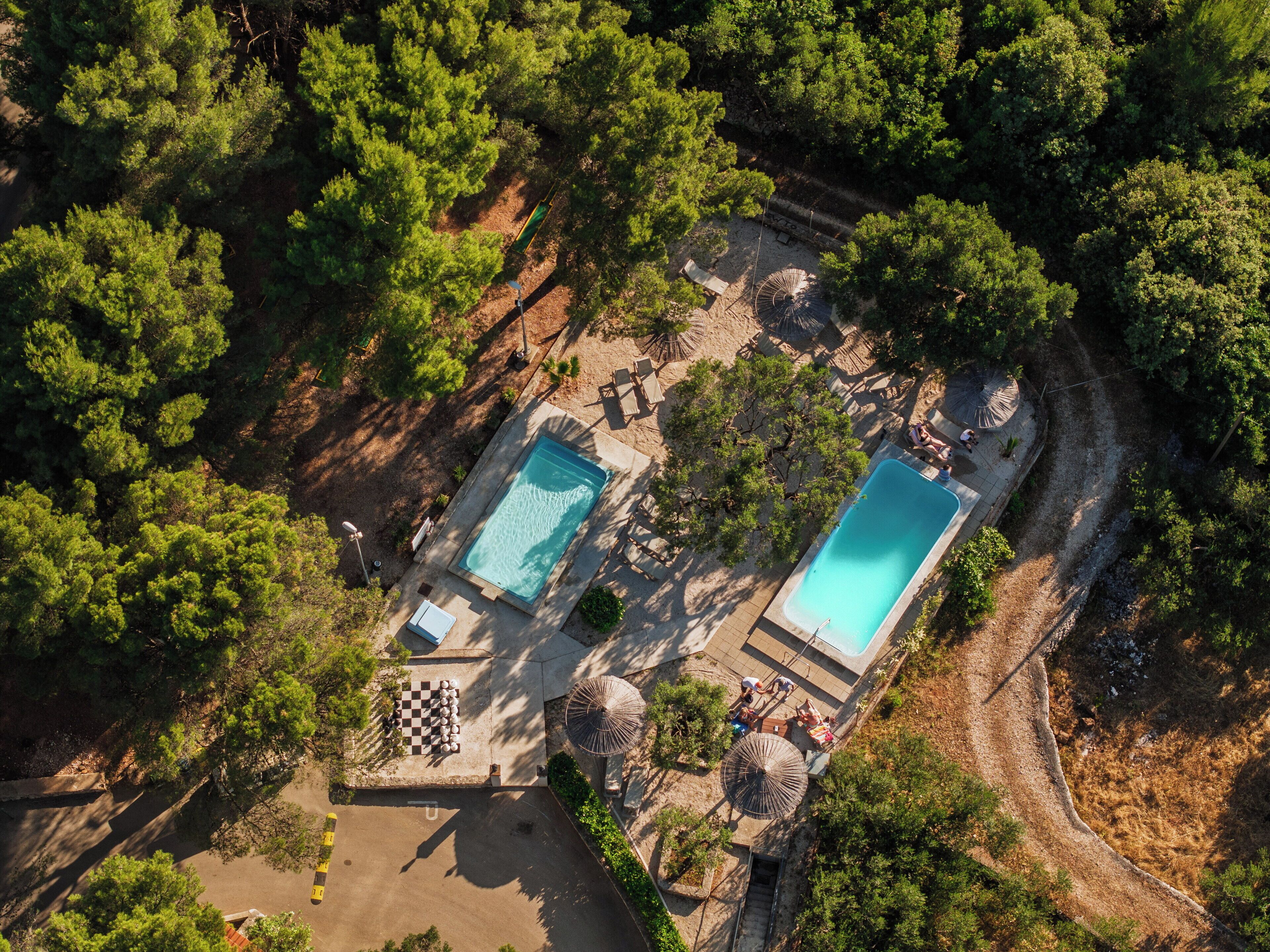 Oversigtsbillede af et resort med to swimmingpools omgivet af træer, et skakternet område og udendørs siddepladser.