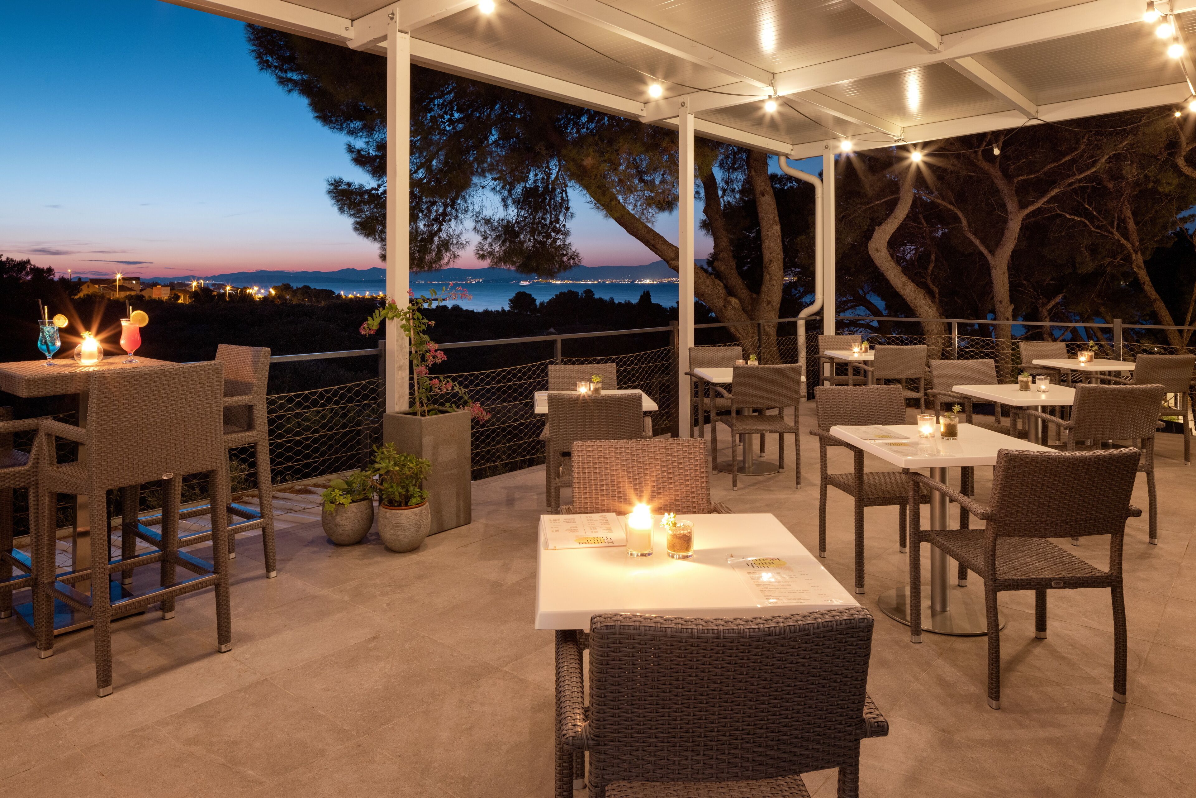 An outdoor patio with tables and chairs, overlooking a scenic view of the sea and mountains at dusk.