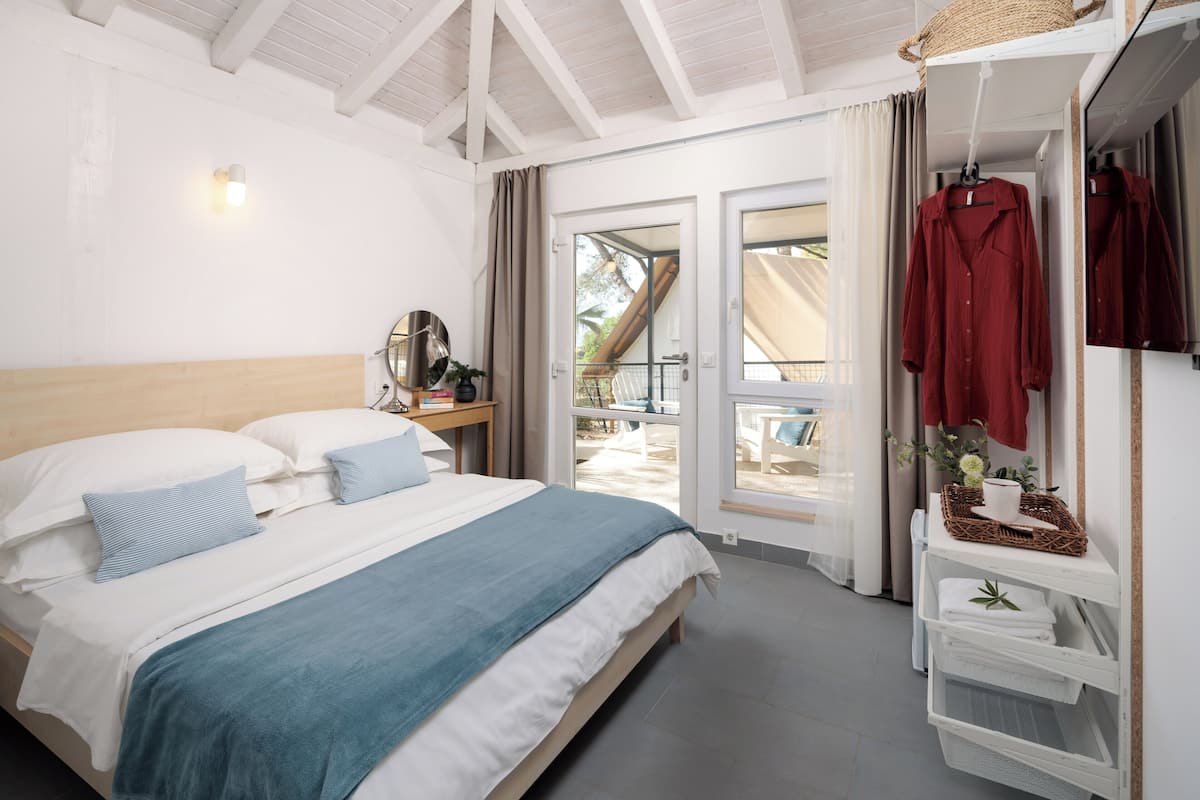 A bedroom with a bed, a nightstand, a mirror, a wardrobe, and a view of the outside.