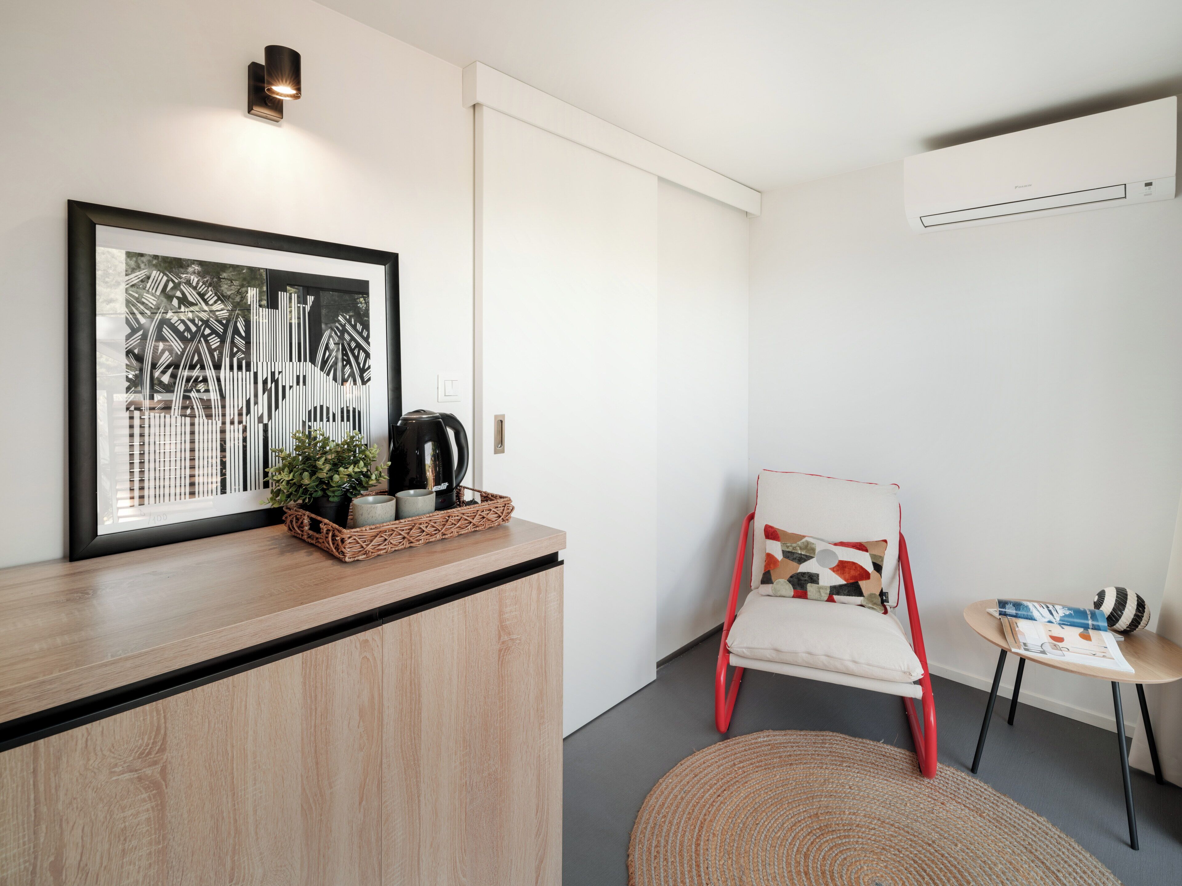 A modern room with a wooden counter, a framed cityscape artwork, a black coffee maker, a red chair, a small round table, and a woven rug.